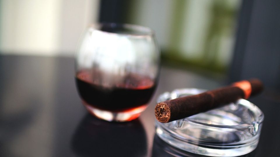 Cigarro puro en un cenicero junto a un vaso con alcohol.
