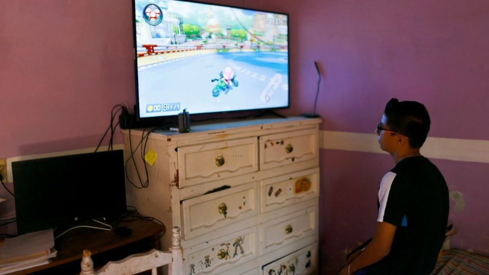 Un joven juega videojuegos en un televisor nuevo enviado por su padre, quien trabaja en EU.