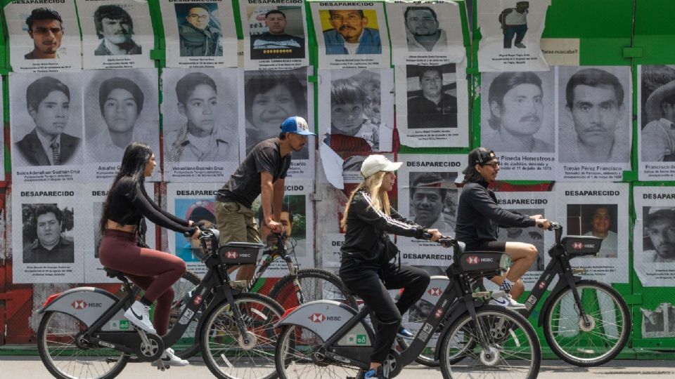 Ciclistas pasan frente a los rostros de personas reportadas como desaparecidas.