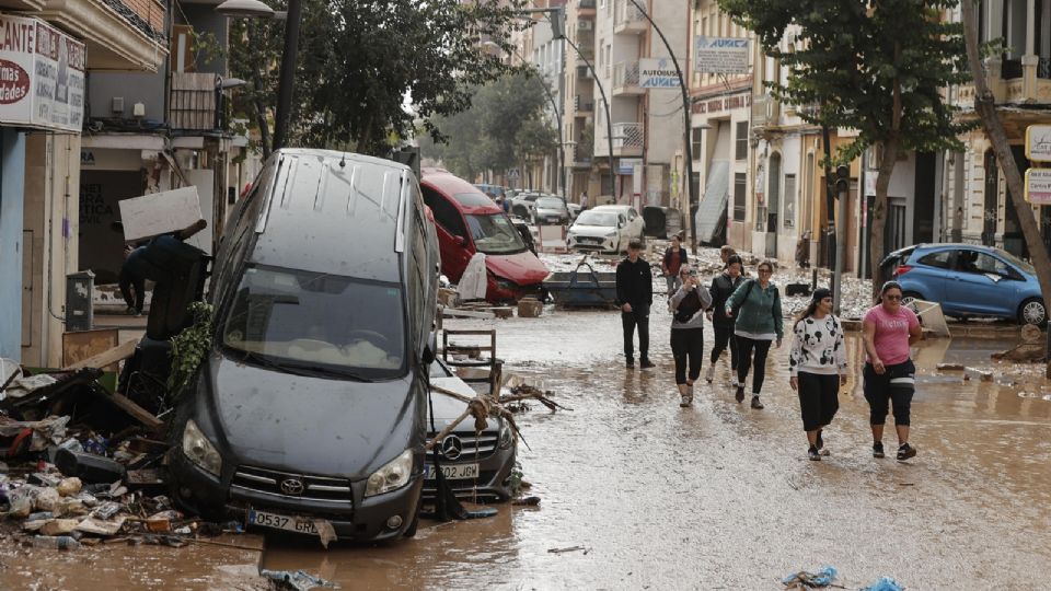 Unas personas caminan por una calle cubierta de lodo y llena de coches amontonados tras las intensas lluvias por la fuerte dana en Valencia.