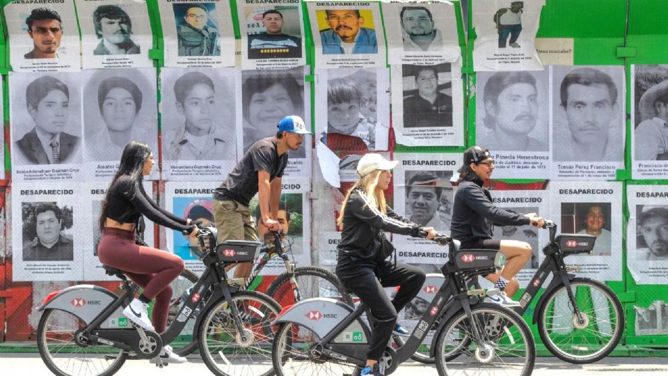 Ciclistas pasan frente a los rostros de personas reportadas como desaparecidas en la Glorieta de las Desaparecidas y Desaparecidos.